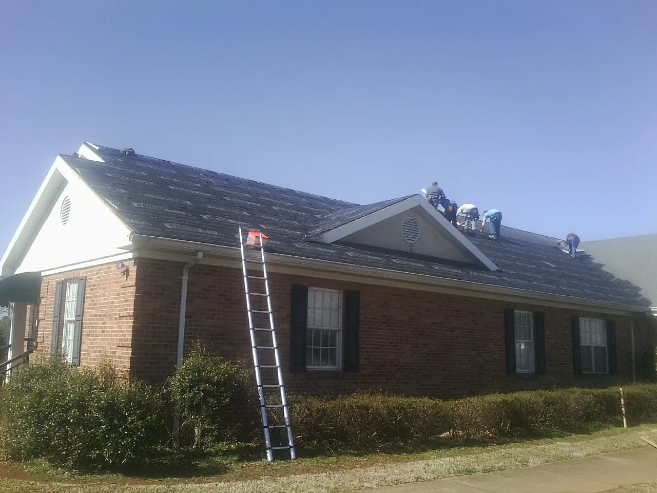 Crew tearing shingles off a brick ranch roof with a ladder set — Emergency Roof Repair work by our crew in Colrain