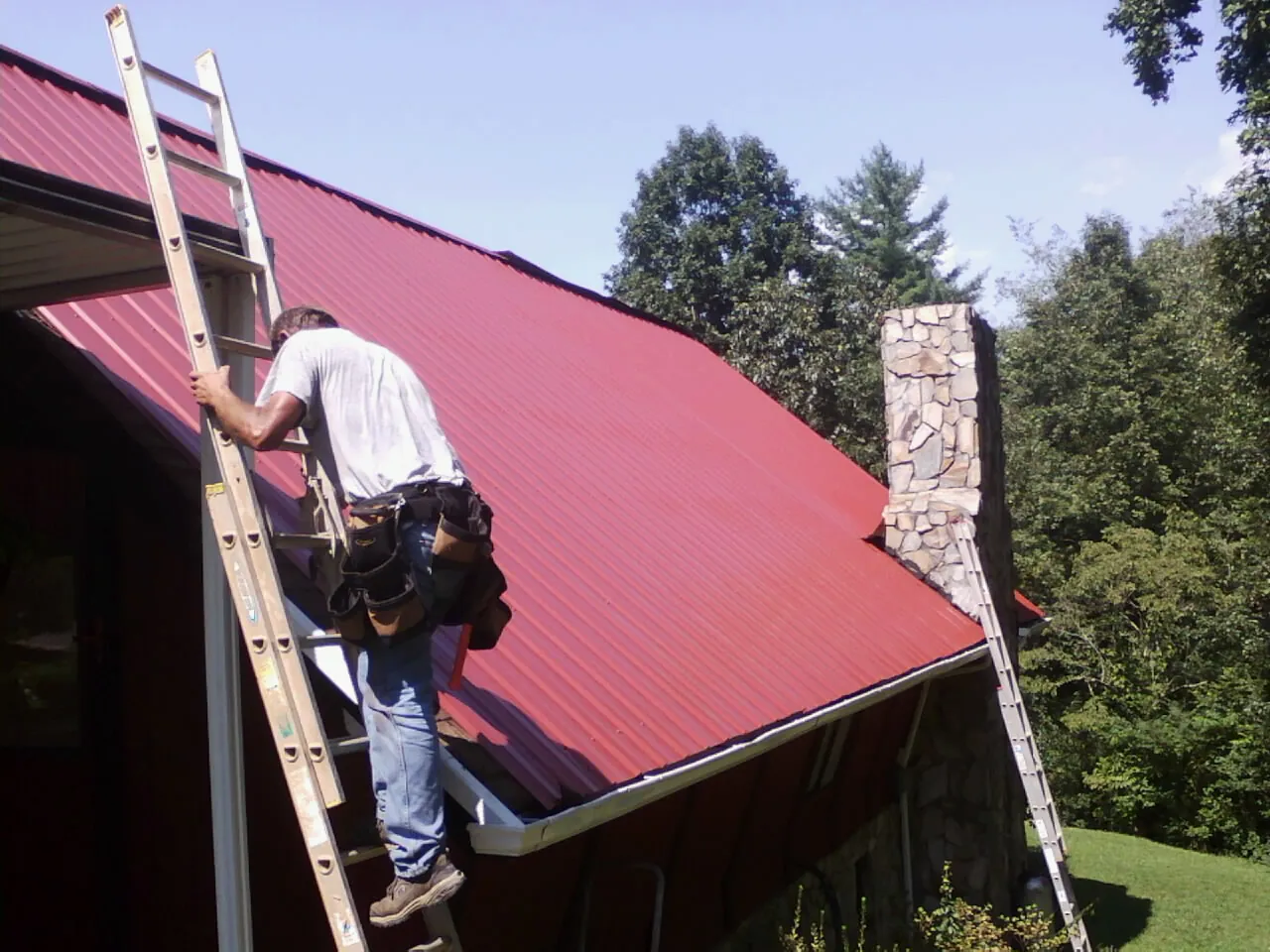 Roofer on a ladder beside a red metal roof and stone chimney — Soffit & Fascia Repair by Roofing Brothers in Colrain